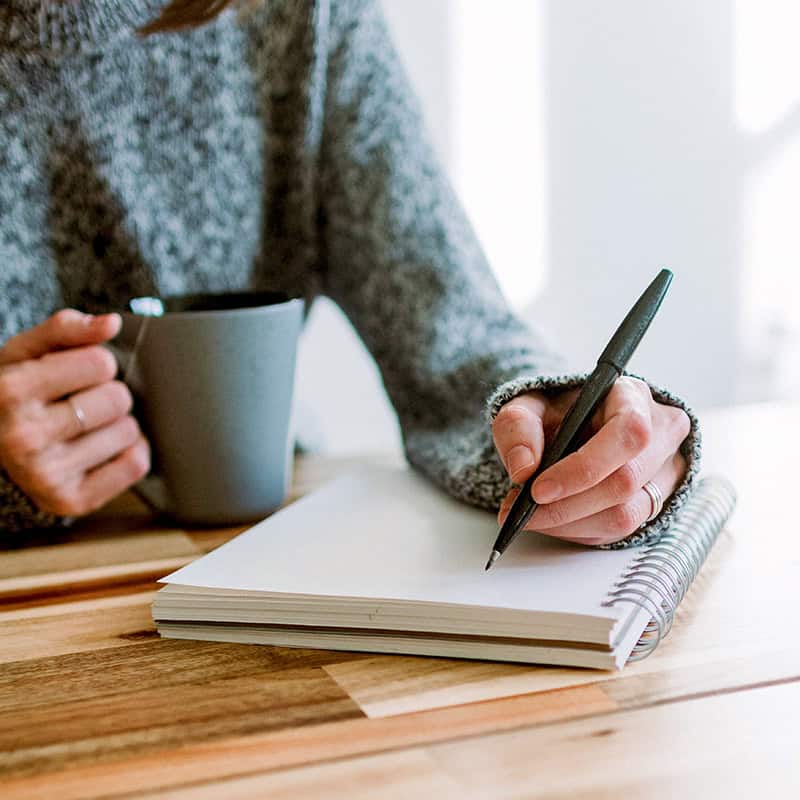 A woman with a warm drink, a notebook, and a pen