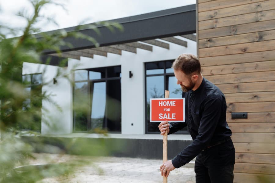 A man putting a sign up in front of a beautiful house. Sign only says Home for Sale with no other details