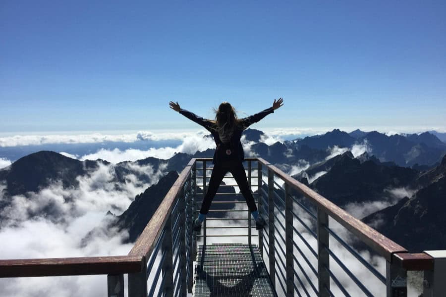 Woman standing on a metal lookout platform overlooking mountain tops.